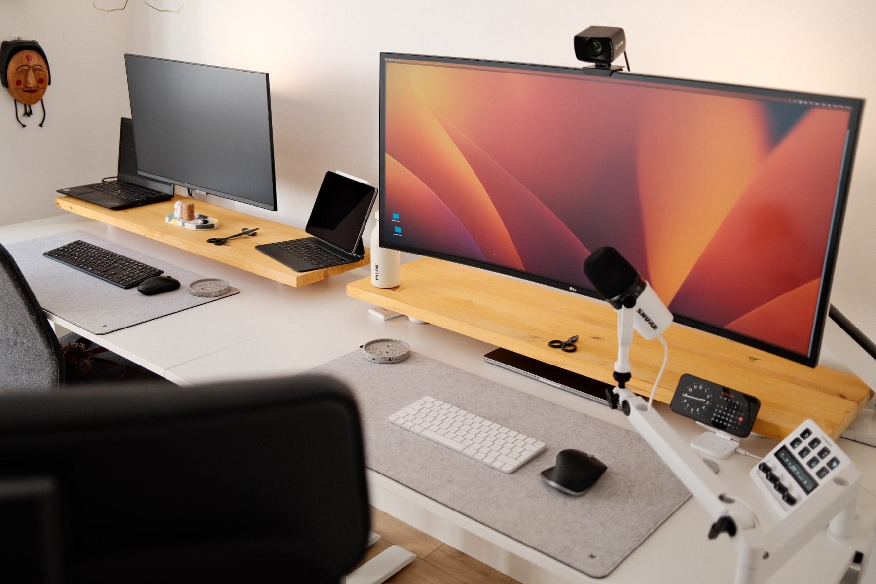 A neatly arranged shared workstation with two monitors, a laptop on a riser, a boom microphone, and assorted peripherals on a wooden desk, with a black office chair partially visible