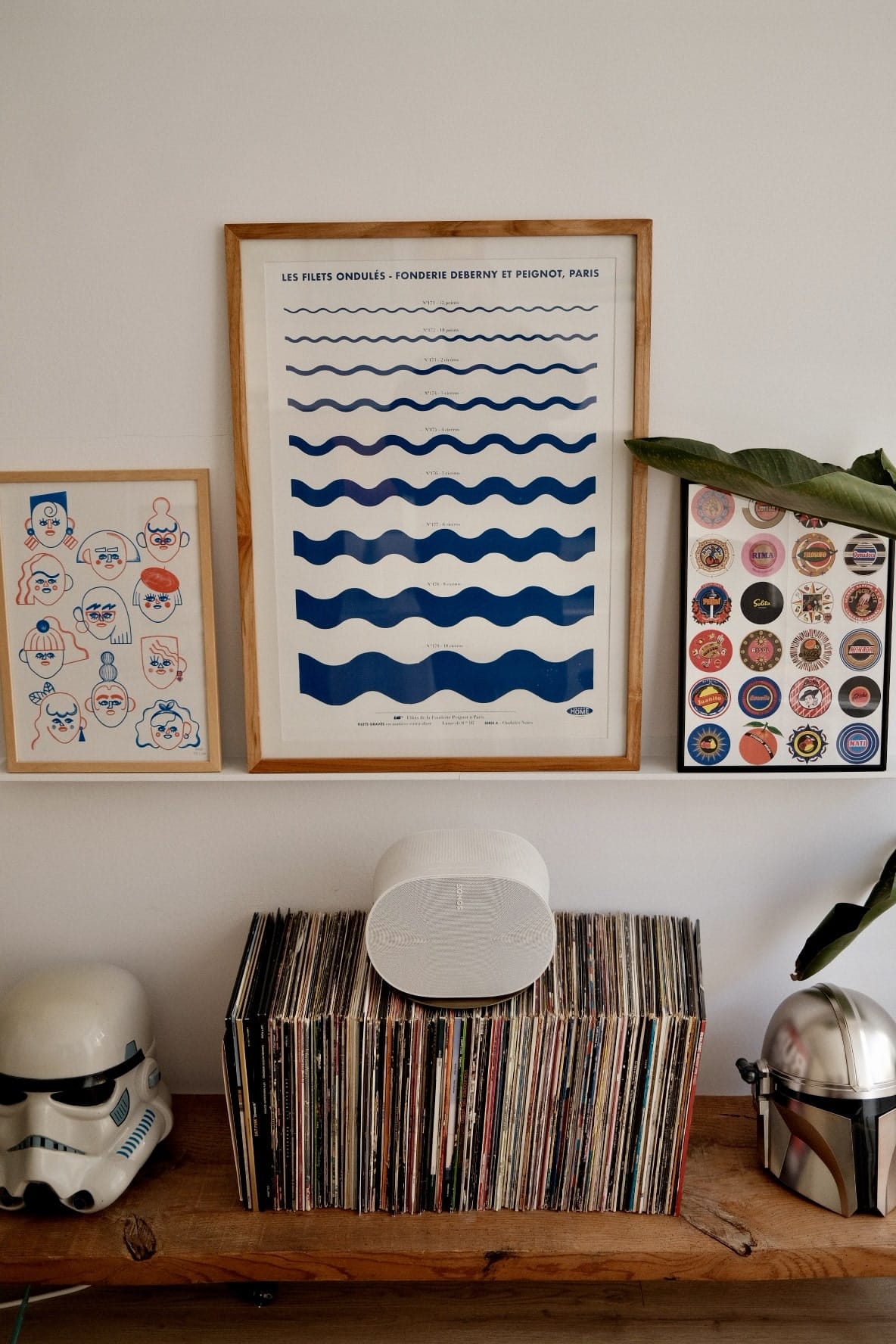 A wooden shelf displaying a collection of vinyl records, flanked by a helmet on each side, with framed graphic art prints hanging above