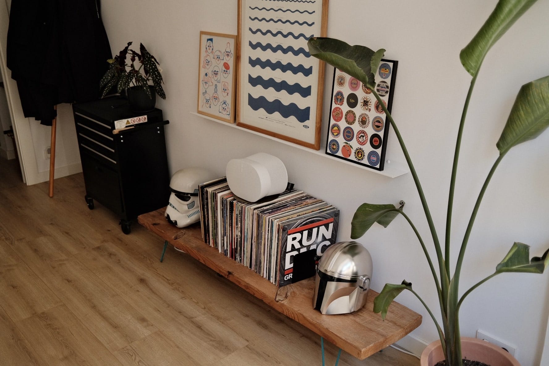 A corner of a room with a collection of vinyl records on a wooden shelf, framed artwork on the wall, a black storage trolley, and a large potted plant