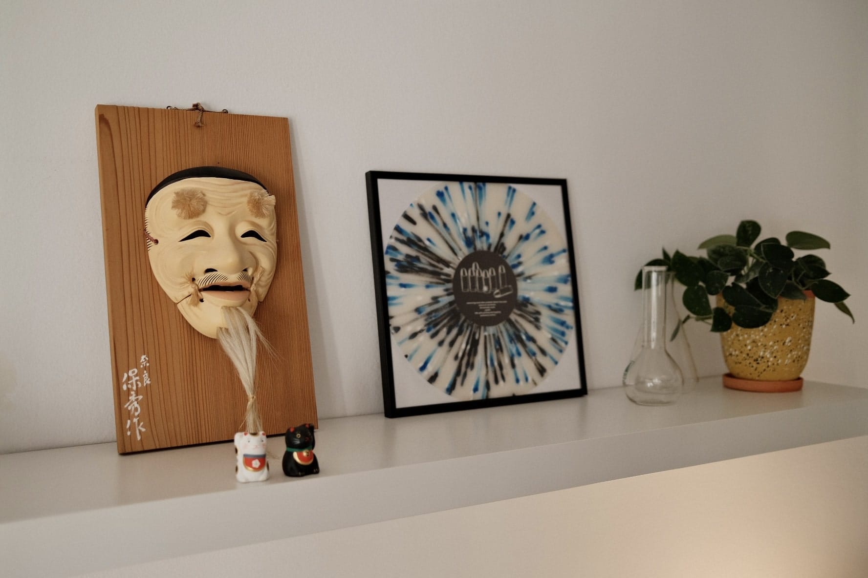 A shelf displaying a traditional wooden mask, two miniature figurines, a framed vinyl record design, and a potted plant in a speckled pot
