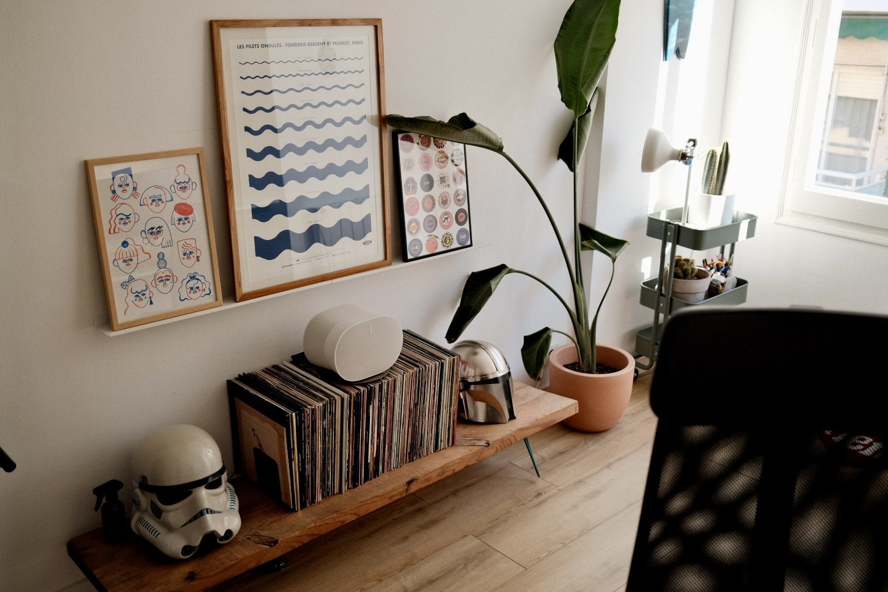 A room corner with a vinyl record collection on a wooden shelf, framed graphic prints on the wall, a plant in a pot, and a speaker