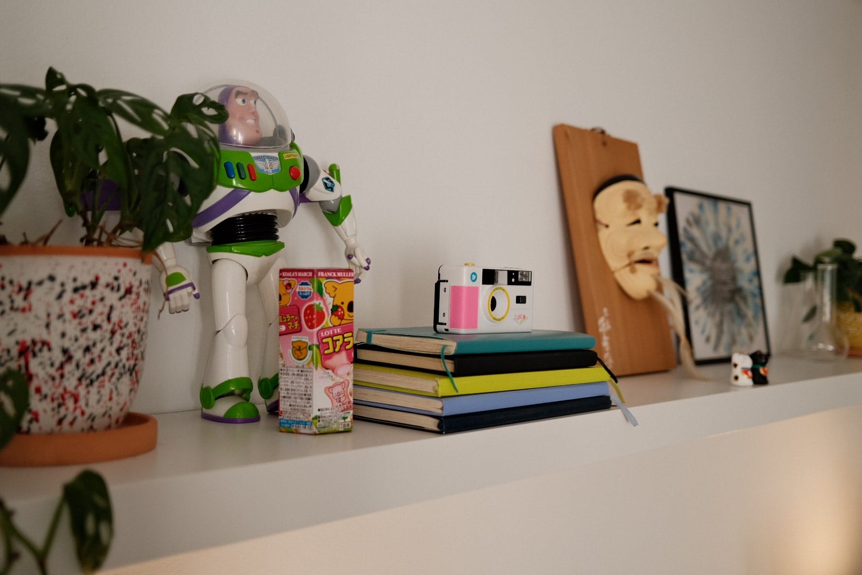 A shelf adorned with an action figure, a potted plant, a colourful instant camera on top of stacked notebooks, and decorative items including a wooden mask and framed artwork