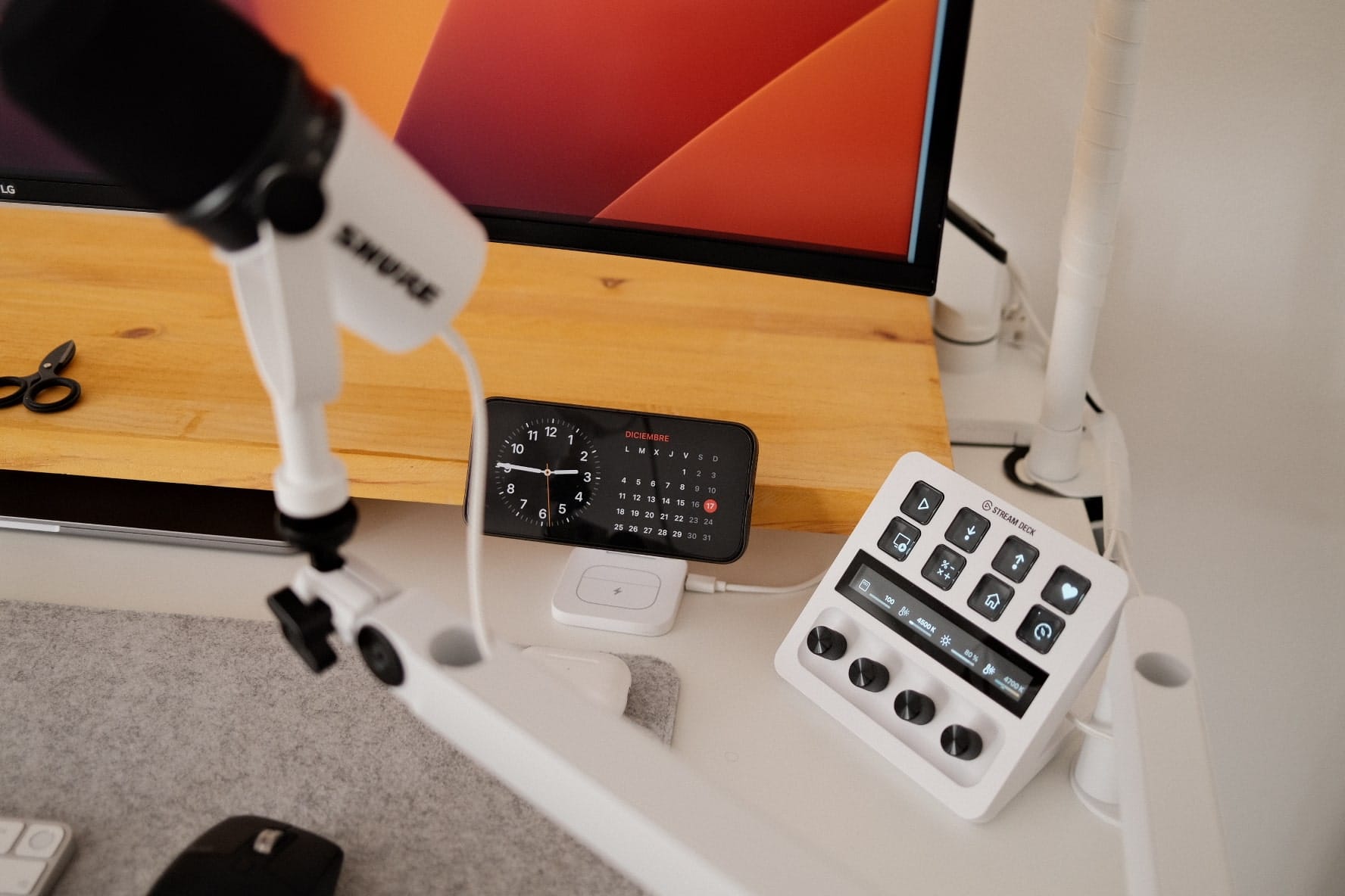 A close-up of a desk with a microphone on a boom arm, a digital clock, and a stream deck next to a computer monitor on a wooden stand