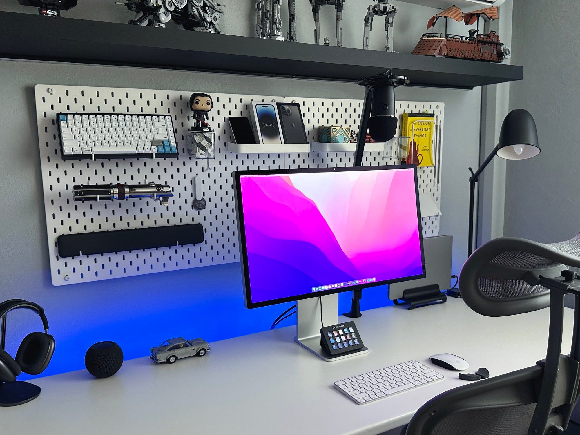 An organised workspace with a monitor, a keyboard, a stream deck, a microphone, a smartwatch, wireless earbuds, a model car, and a book titled The Design of Everyday Things on a shelf above