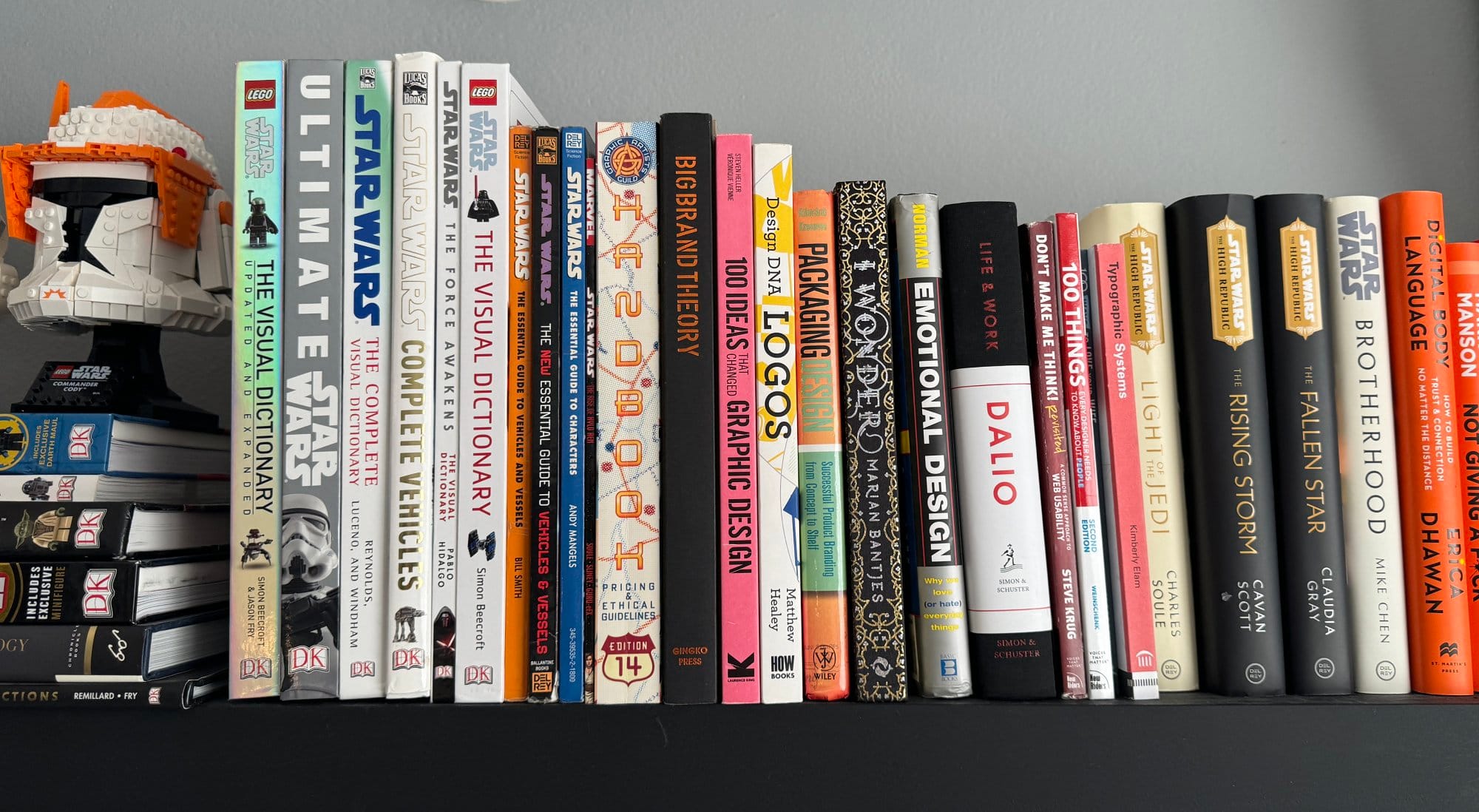 A shelf holding a variety of books primarily focused on Star Wars, design, and graphic arts, along with a LEGO Star Wars helmet model to the left