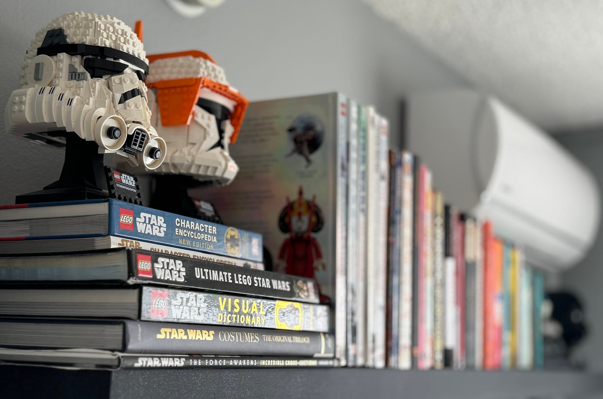 A bookshelf featuring a selection of Star Wars books and encyclopedias, with LEGO Star Wars helmet models displayed on top, and other books blurred in the background