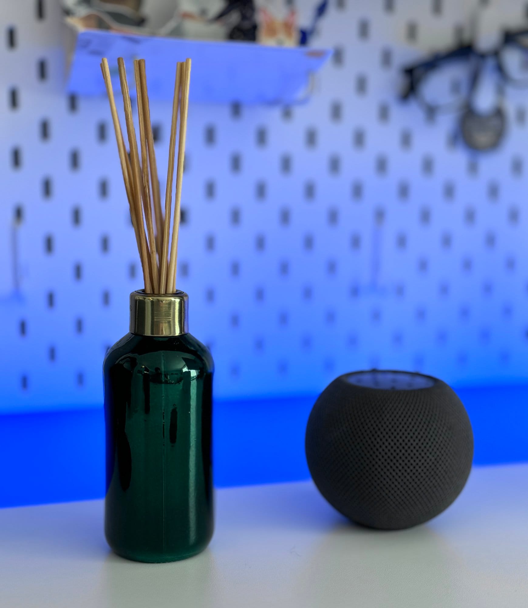 A green reed diffuser bottle with sticks on a white surface, with a blurred background featuring a pegboard and a black speaker