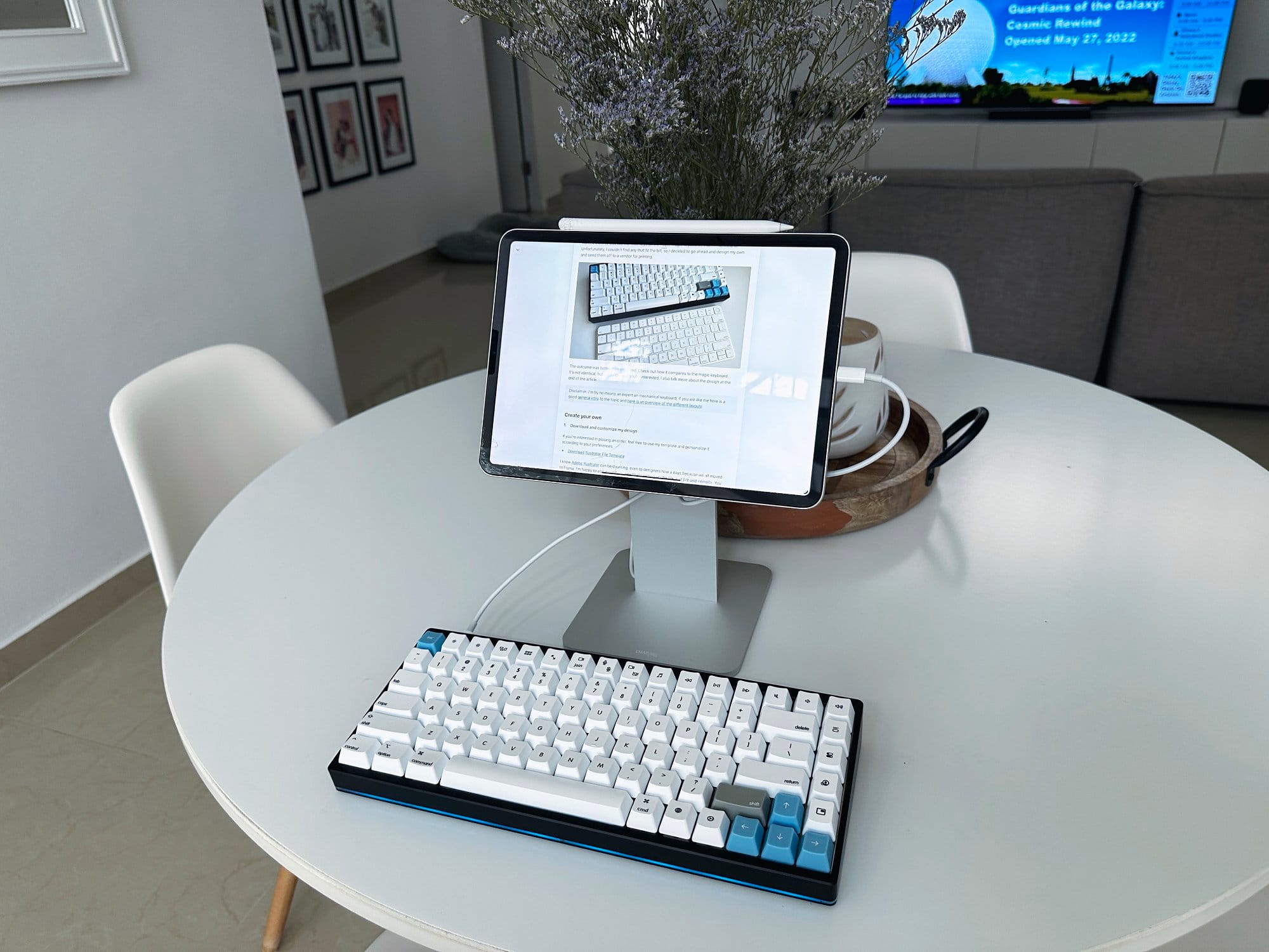 A modern minimalist workspace setup with a tablet mounted on a stand, paired with a mechanical keyboard on a white round table, and a cosy living area in the background