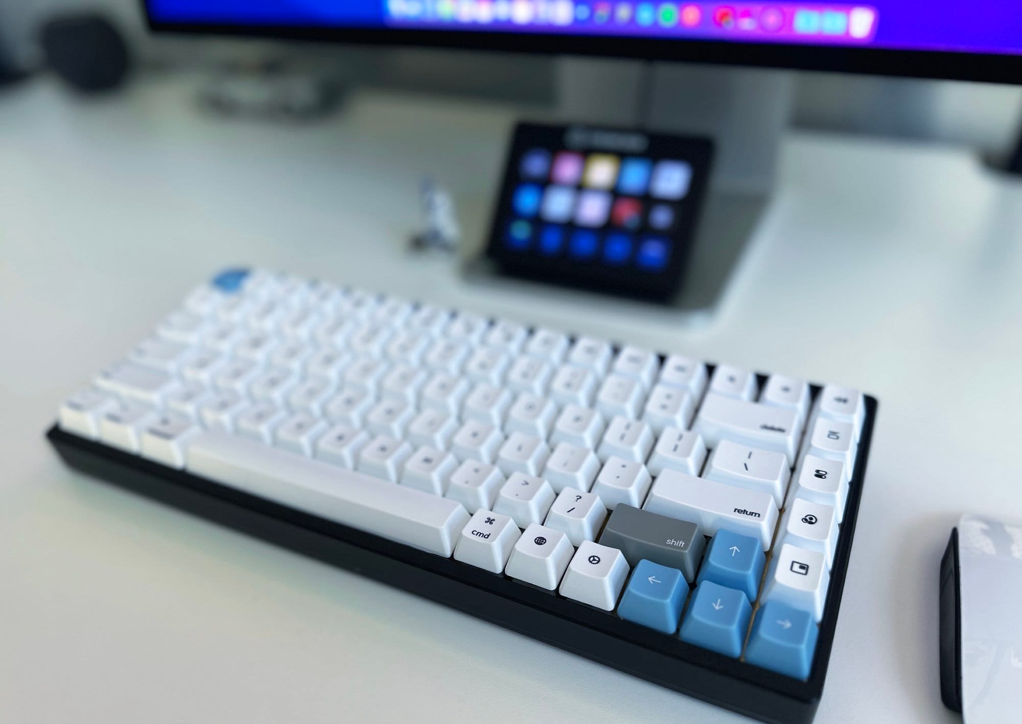 A close-up of a mechanical keyboard on a desk, with blurred background elements including a mouse, a tablet on a stand, and part of a monitor