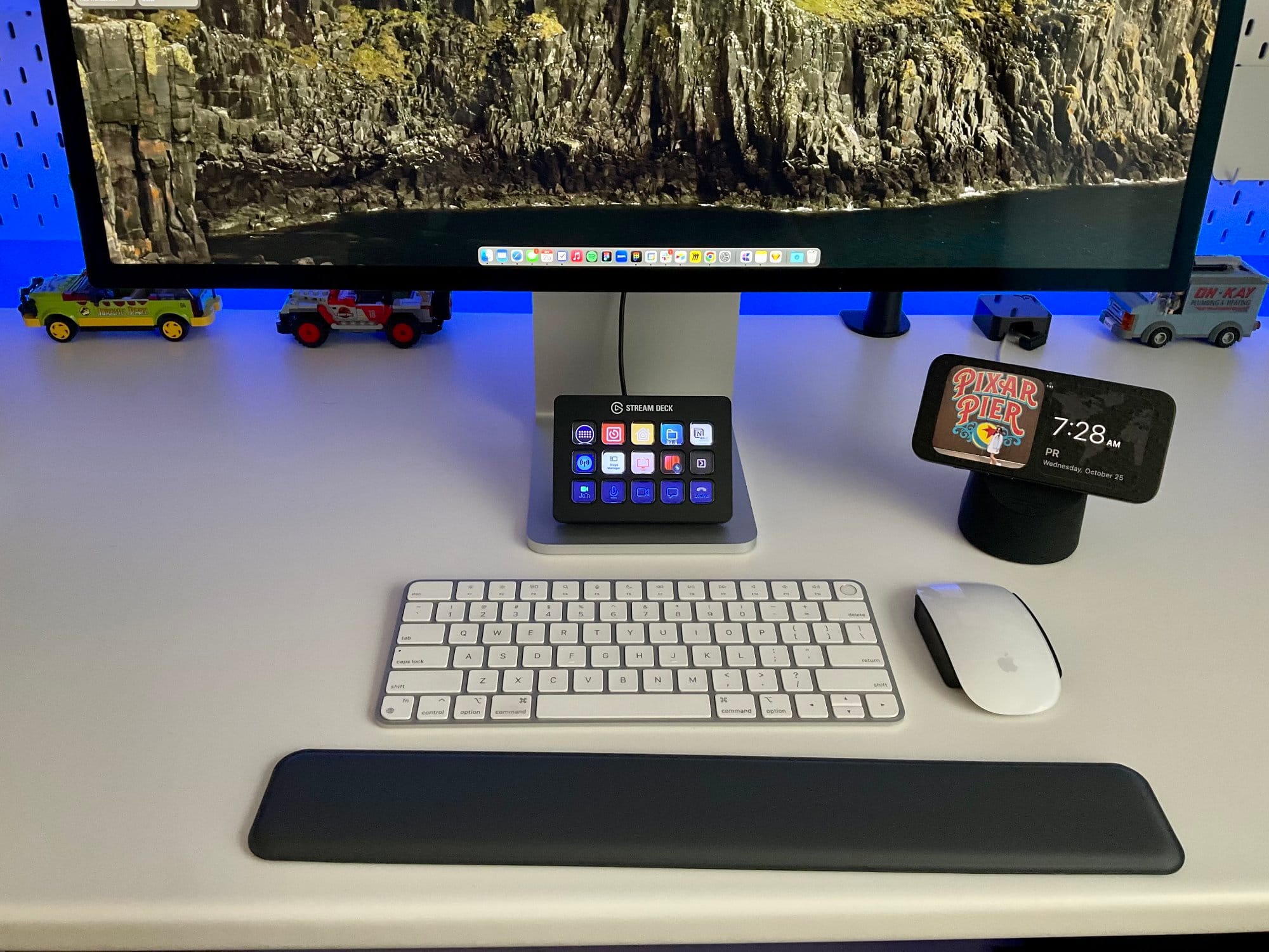 A desk with a monitor, keyboard, mouse, a stream deck with icons, a smartphone showing the time and a weather widget, and two toy cars underneath the monitor