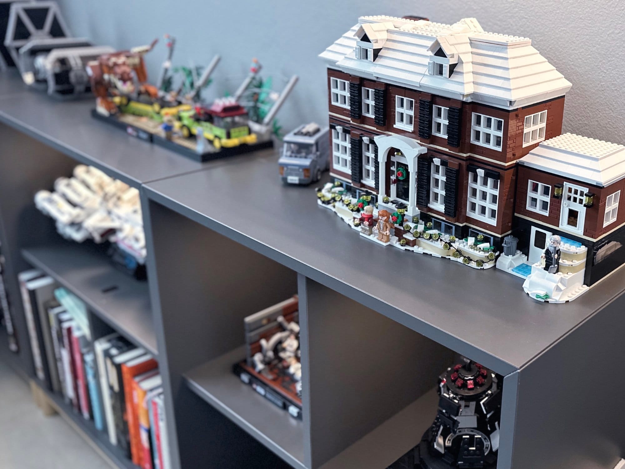 A shelf displaying a collection of intricate models, including a detailed brick-built house and various vehicles, with books stored on the lower shelf
