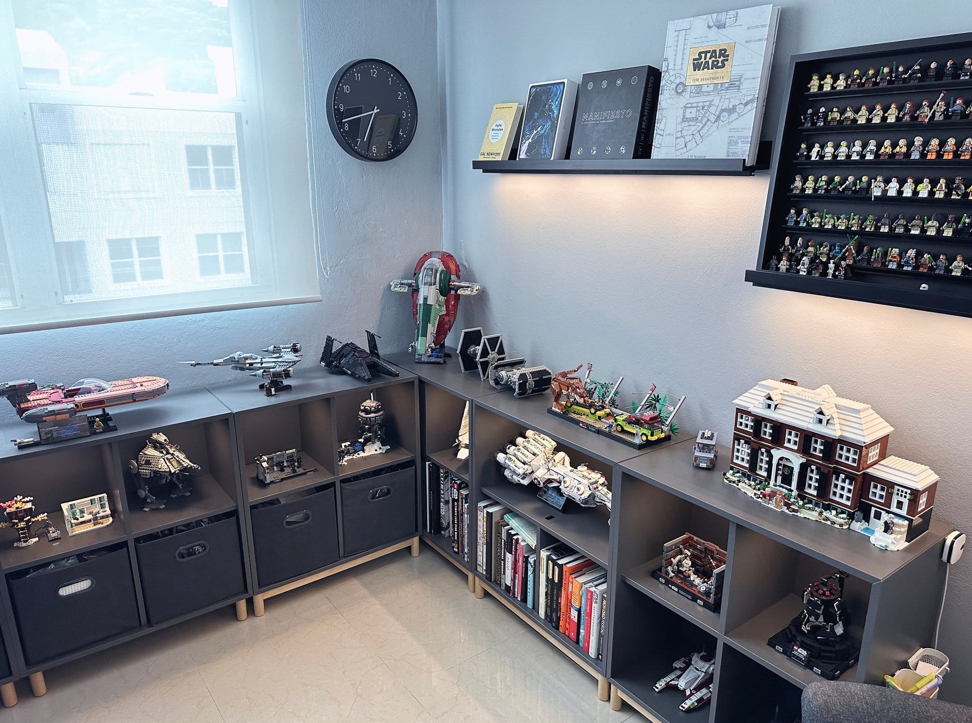 A room corner featuring a shelving unit with various assembled models and vehicles on display, a collection of action figures, and a selection of books, with a clock and framed pictures on the wall above