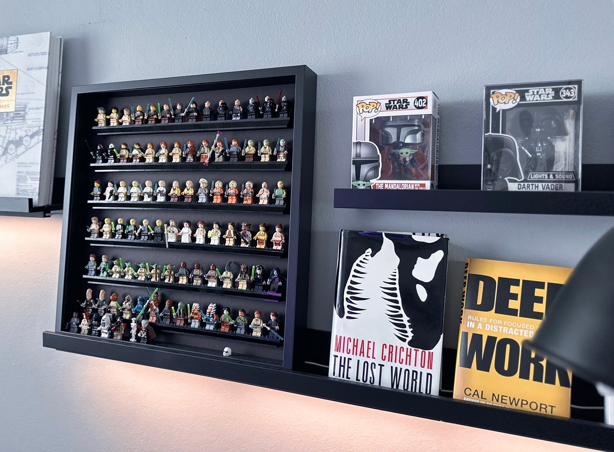 On the wall shelf, there are rows of collectible figures, two boxed figurines, and books including The Lost World by Michael Crichton and Deep Work by Cal Newport