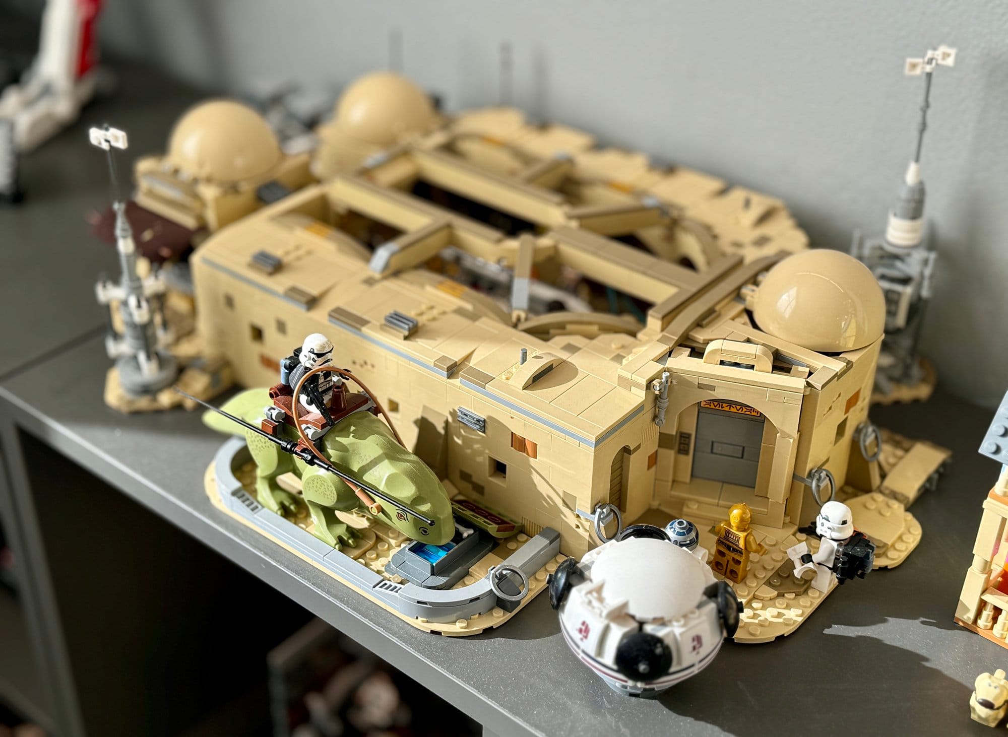 A detailed LEGO 75290 Star Wars model scene with a central tan building, a green and brown speeder with a figure, and various characters including white-armoured figures around it