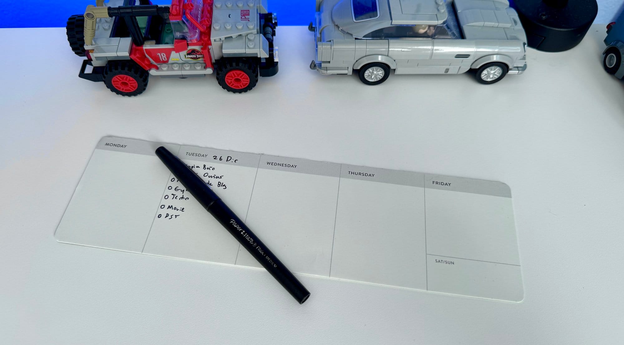 A desk with a weekly planner pad and a pen, some written tasks for Tuesday, and two model vehicles on either side, against a backdrop of a work environment