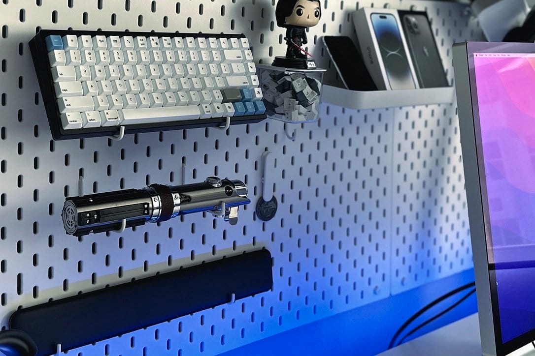A pegboard organising a mechanical keyboard, a replica lightsaber, a bobblehead figure, and VR controllers, with a glimpse of a monitor on the right
