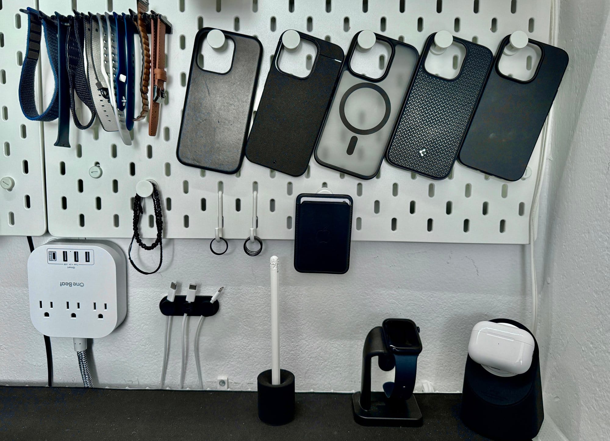 A pegboard organising space with various phone cases hung up, charging cables, a smartwatch on a stand, wireless earbuds in a case, and a multi-port power outlet on the left