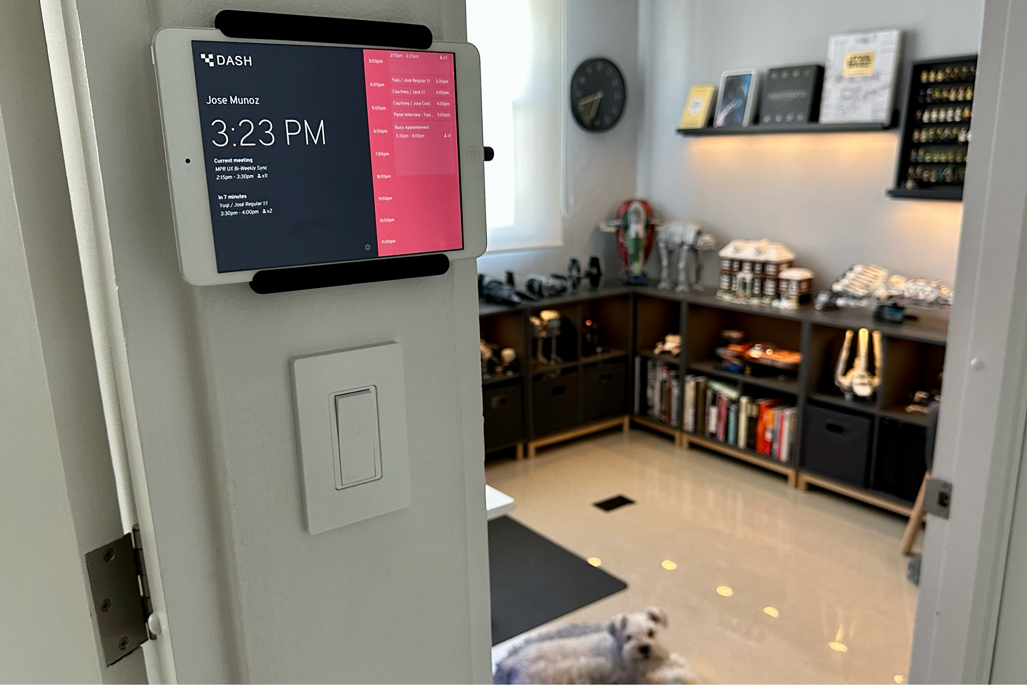 A door-mounted tablet displaying a digital clock and calendar schedule, with a blurred background showing a room with shelves, collectibles, books, and a dog lying on the floor