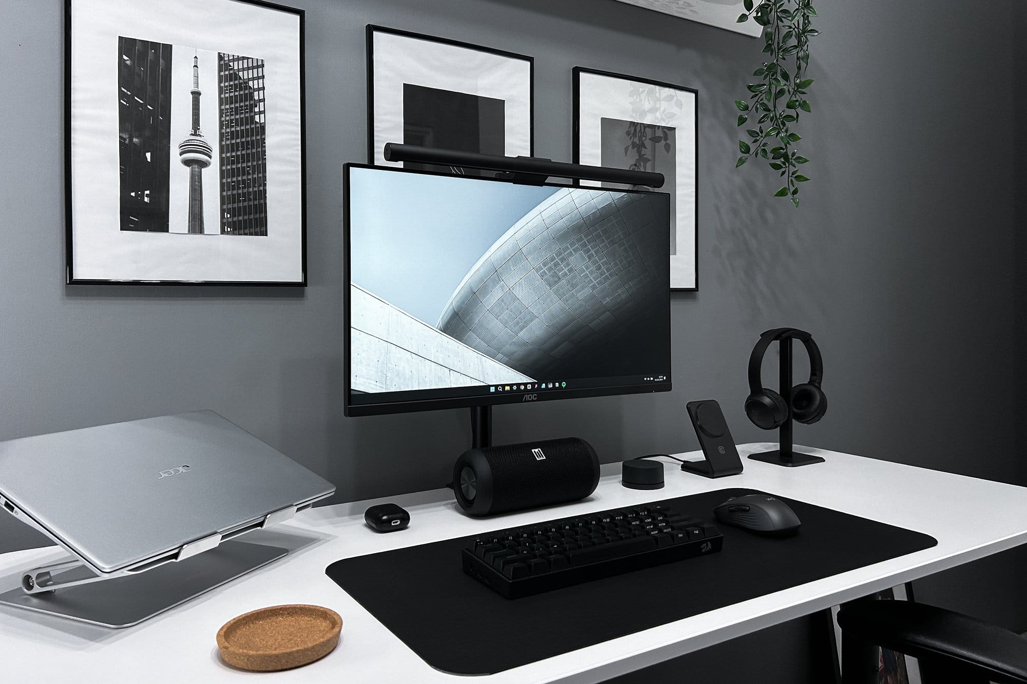 A sleek and modern home office featuring a wide monitor atop a white desk with a black mat, a closed laptop on a stand, a black keyboard, and a wireless mouse