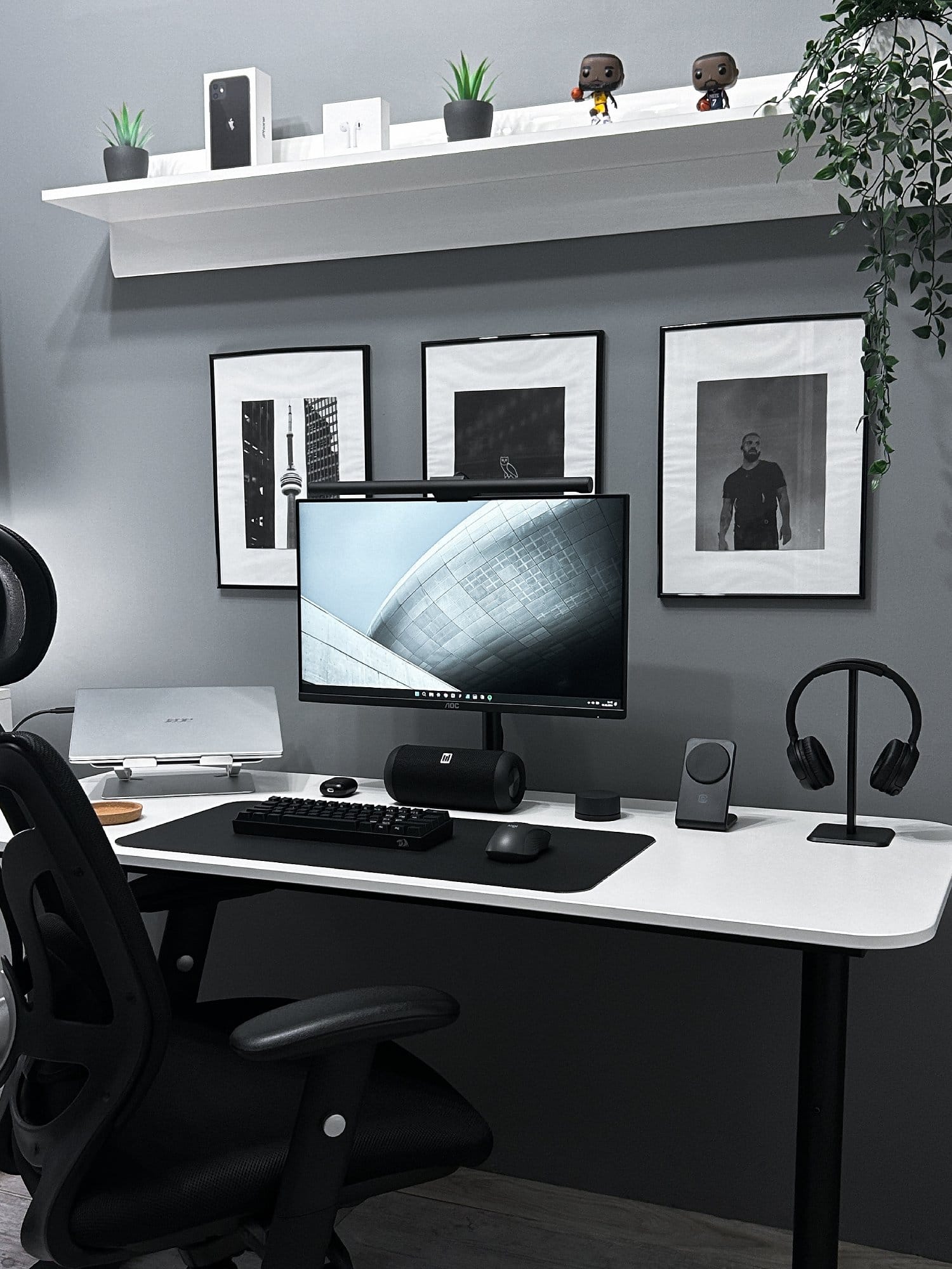 A well-organised desk with a computer monitor, keyboard, and headphones, complemented by decorative plants and figurines on a shelf and framed pictures on the wall in a home office setup