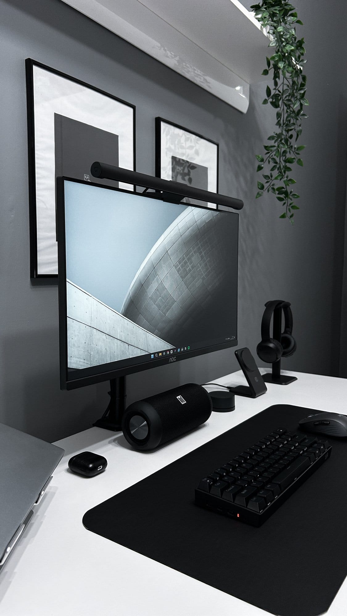 A modern and organised workspace with a large computer monitor, a mechanical keyboard, a cylindrical speaker, and headphones, accented by framed artwork and a hanging plant on a grey wall