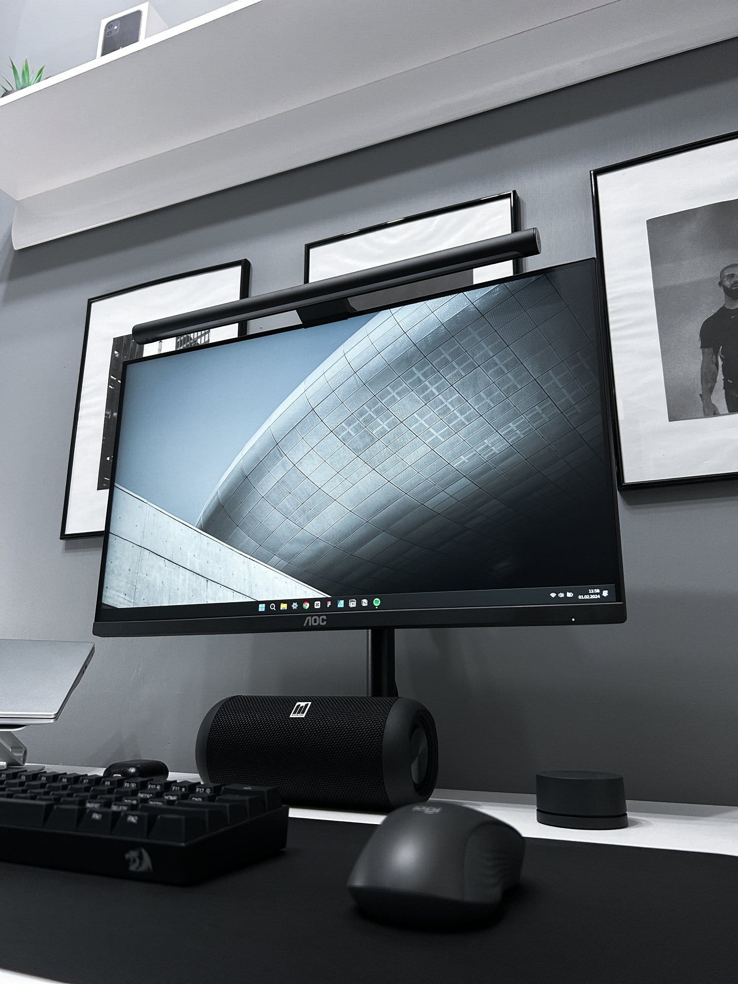 A home office setup with a large monitor, mechanical keyboard, speaker, and mouse on a desk, with monochrome framed pictures on the grey wall above