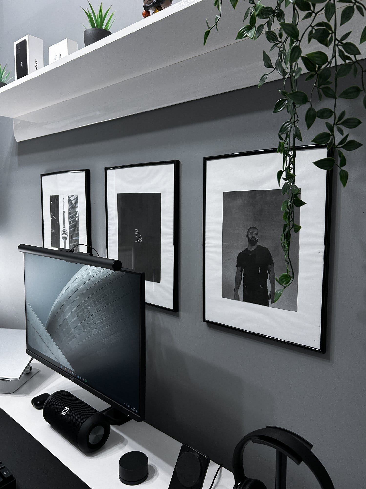 A home office corner with a monitor, speaker, and headphones on a white desk, with framed black and white pictures on the wall, complemented by a trailing plant and decorative items on a shelf above