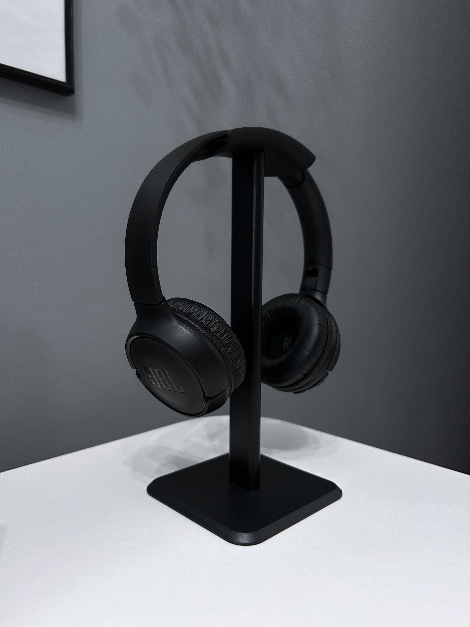 Black over-ear JBL headphones resting on a stand on a white surface against a grey background with a framed picture partially visible