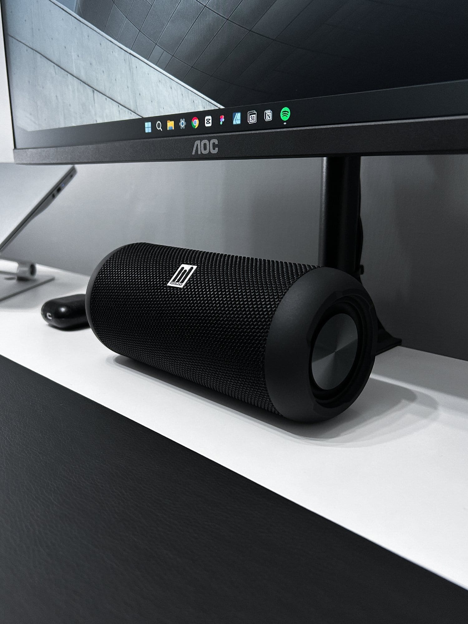 A close-up of a black cylindrical desktop speaker in front of a monitor on a white desk with a black surface