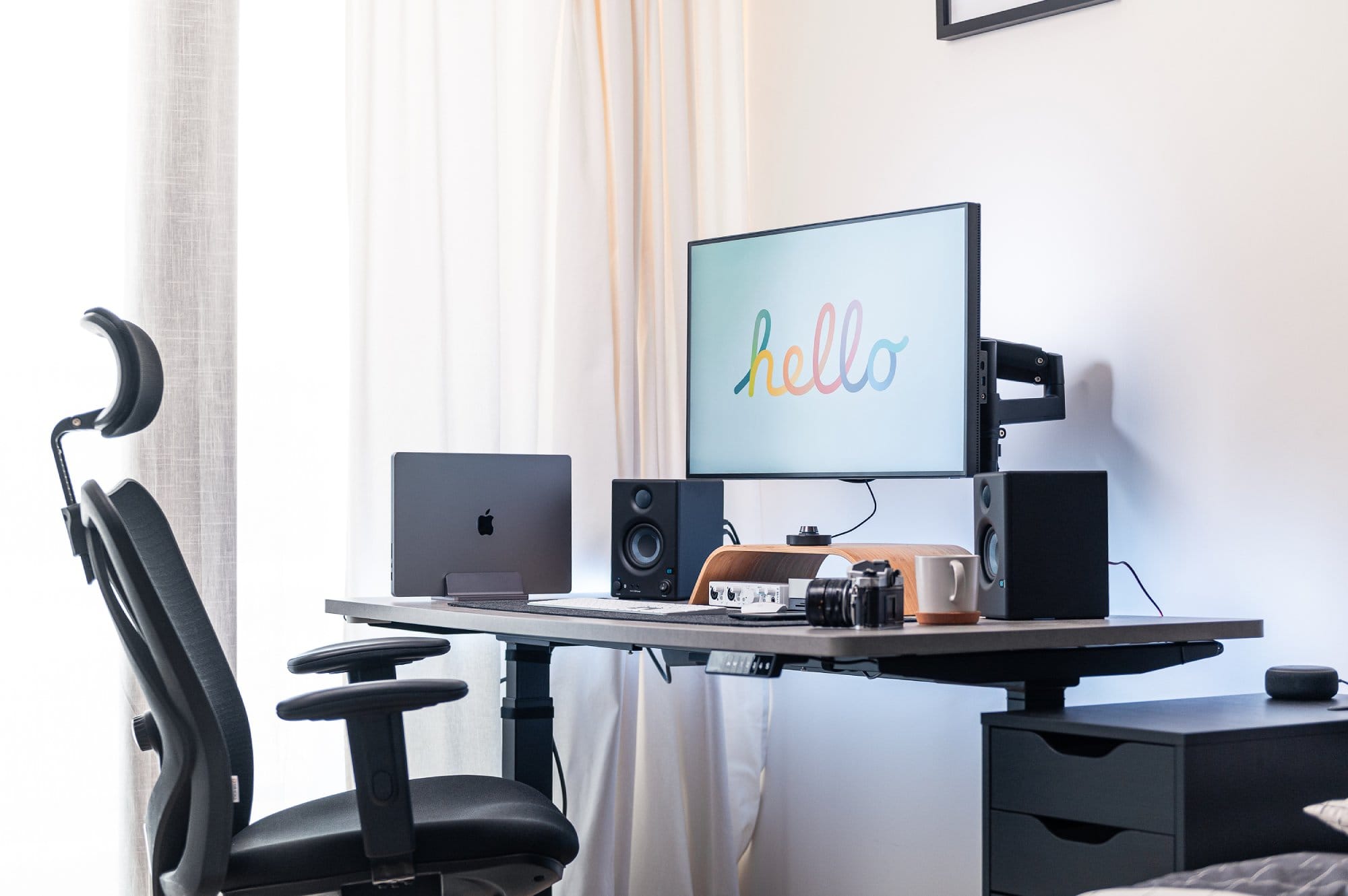 An ergonomic office chair facing a desk with a laptop, a monitor displaying the word “hello” in colourful script, speakers, a camera, and various desk accessories against a window with sheer curtains