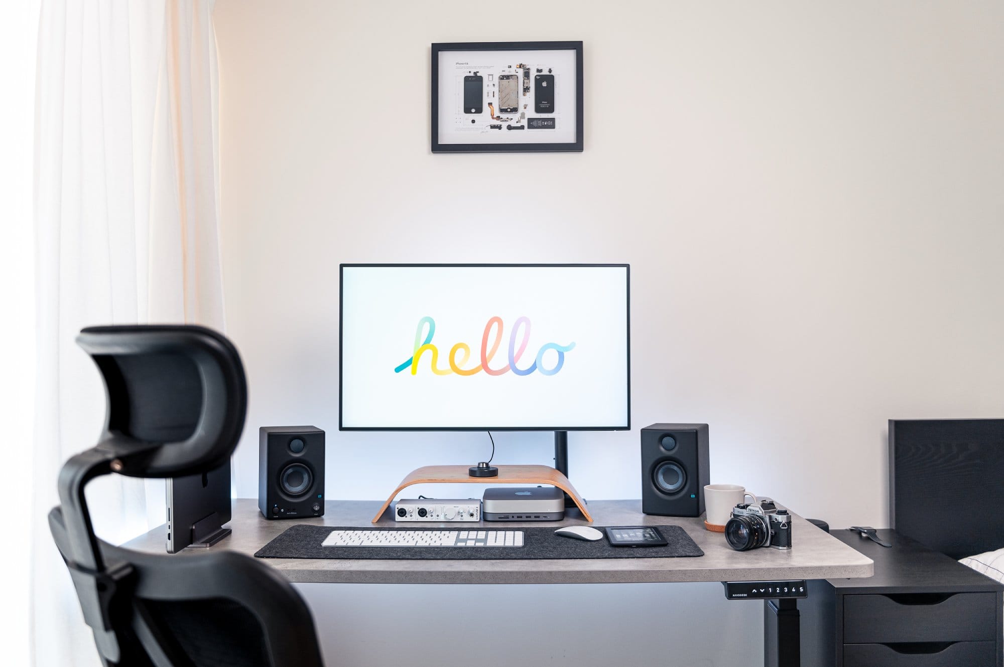 A neatly organised desk space with a central monitor displaying a multicoloured “hello”, speakers on each side, a laptop, camera, and audio equipment, with a framed smartphone schematic on the wall