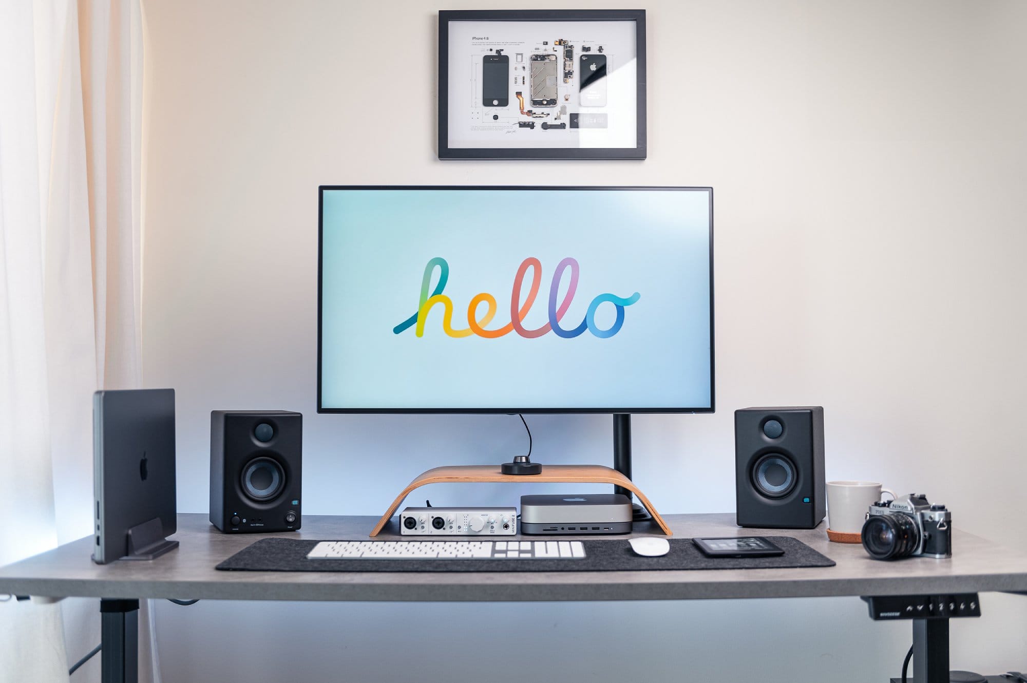A modern desk setup with a monitor displaying the word “hello” in rainbow colours, studio monitors on each side, a laptop on a stand, audio equipment, a white mug, and a camera, with a framed iPhone schematic on the wall