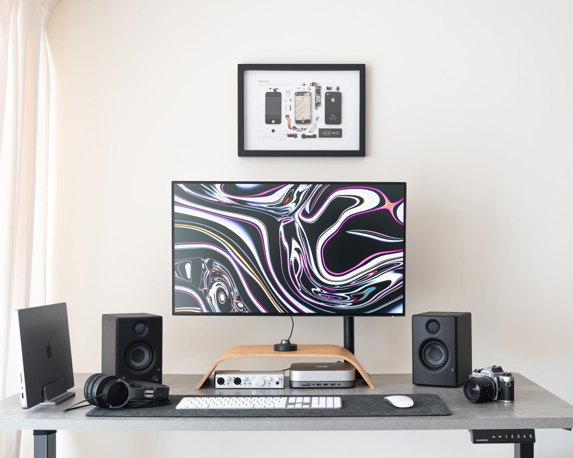 A contemporary desk setup featuring a large monitor with an abstract swirl screensaver, flanked by speakers, with a laptop, headphones, a vintage camera, and a framed diagram of a smartphone on the wall above