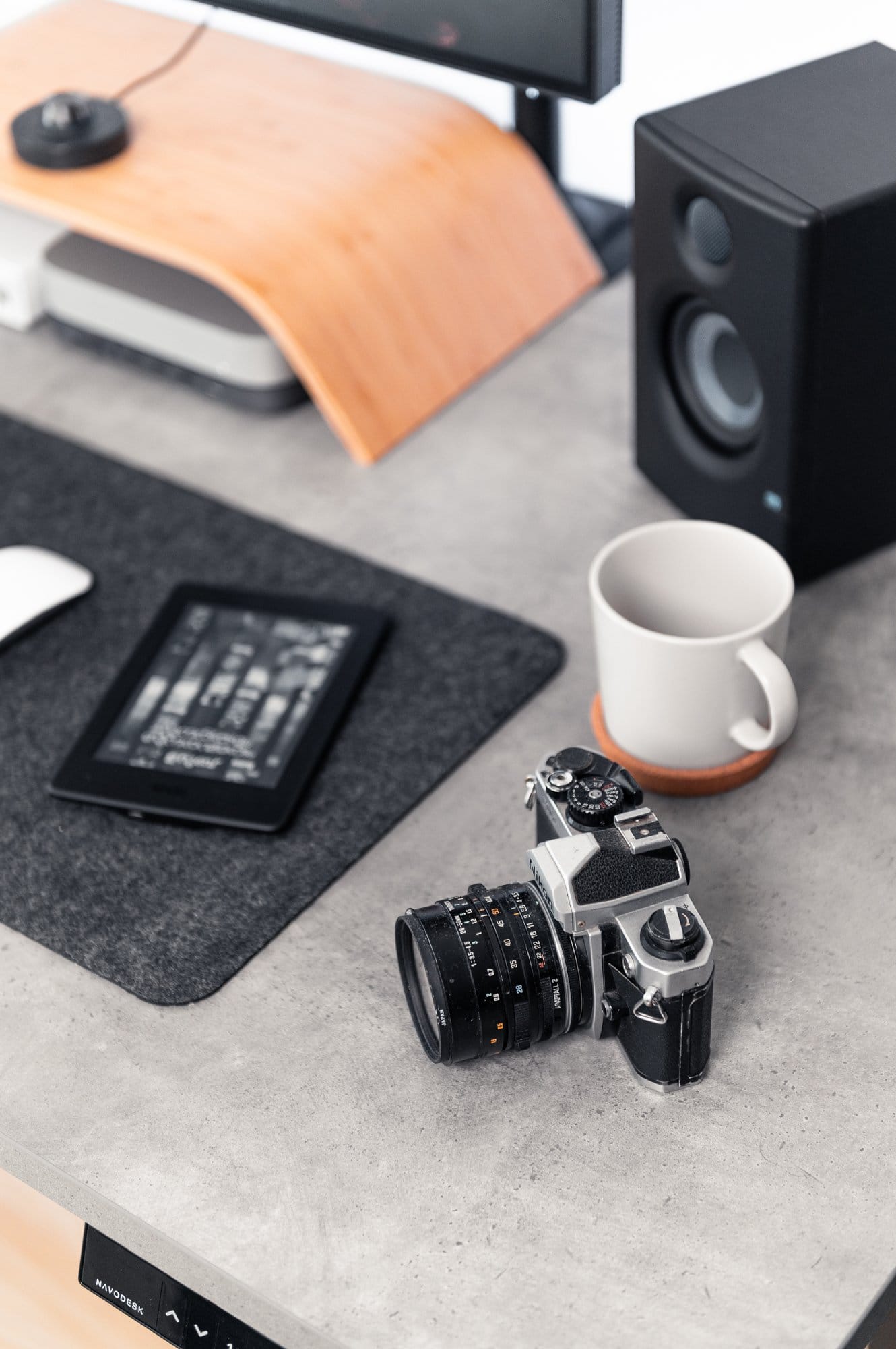 A close-up of a desk with a Nikon FM2 camera, a tablet displaying a book, a white mug on a coaster, and part of a monitor stand and speaker in the background