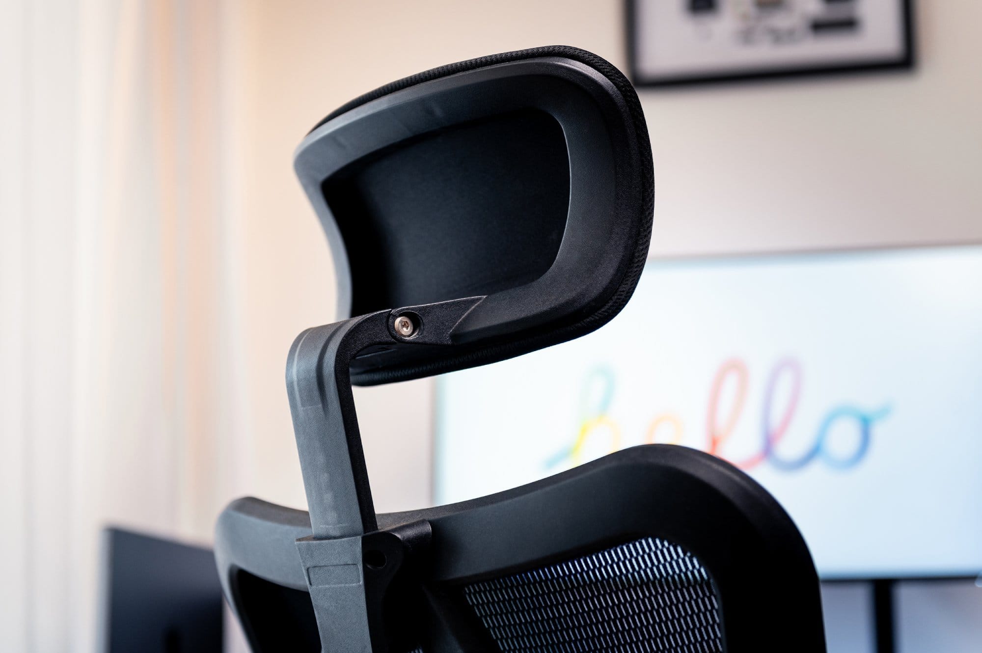 A close-up of the top of a black ergonomic office chair with a blurred background featuring a monitor with colourful text