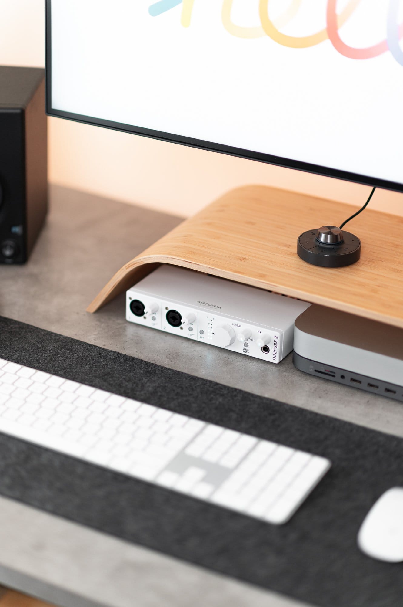 A close-up view of a modern desk with a wooden monitor stand holding an audio interface, next to a laptop, with a part of the keyboard and mouse in the foreground, and a monitor with colourful text in the background