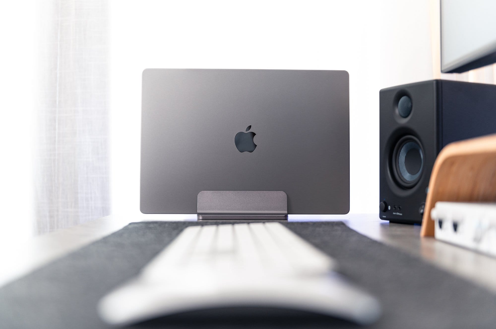 A focused view of a closed MacBook on a stand on a desk, with a blurred foreground showing part of a keyboard and a speaker in the background, all in a bright office setting with a window on the left side