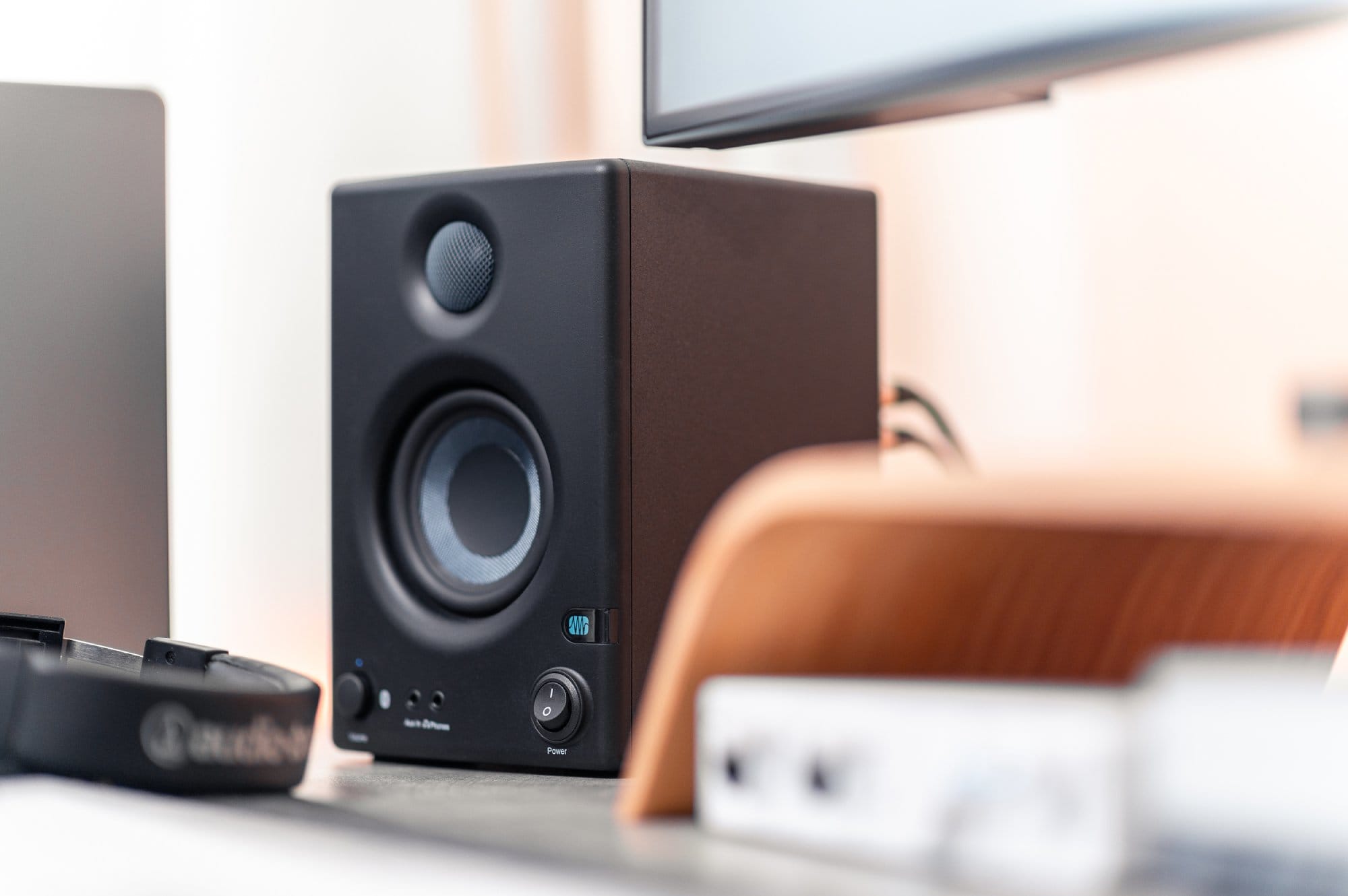 A focused view of a modern black speaker on a desk, with a blurred laptop, monitor stand, and other office equipment in the background