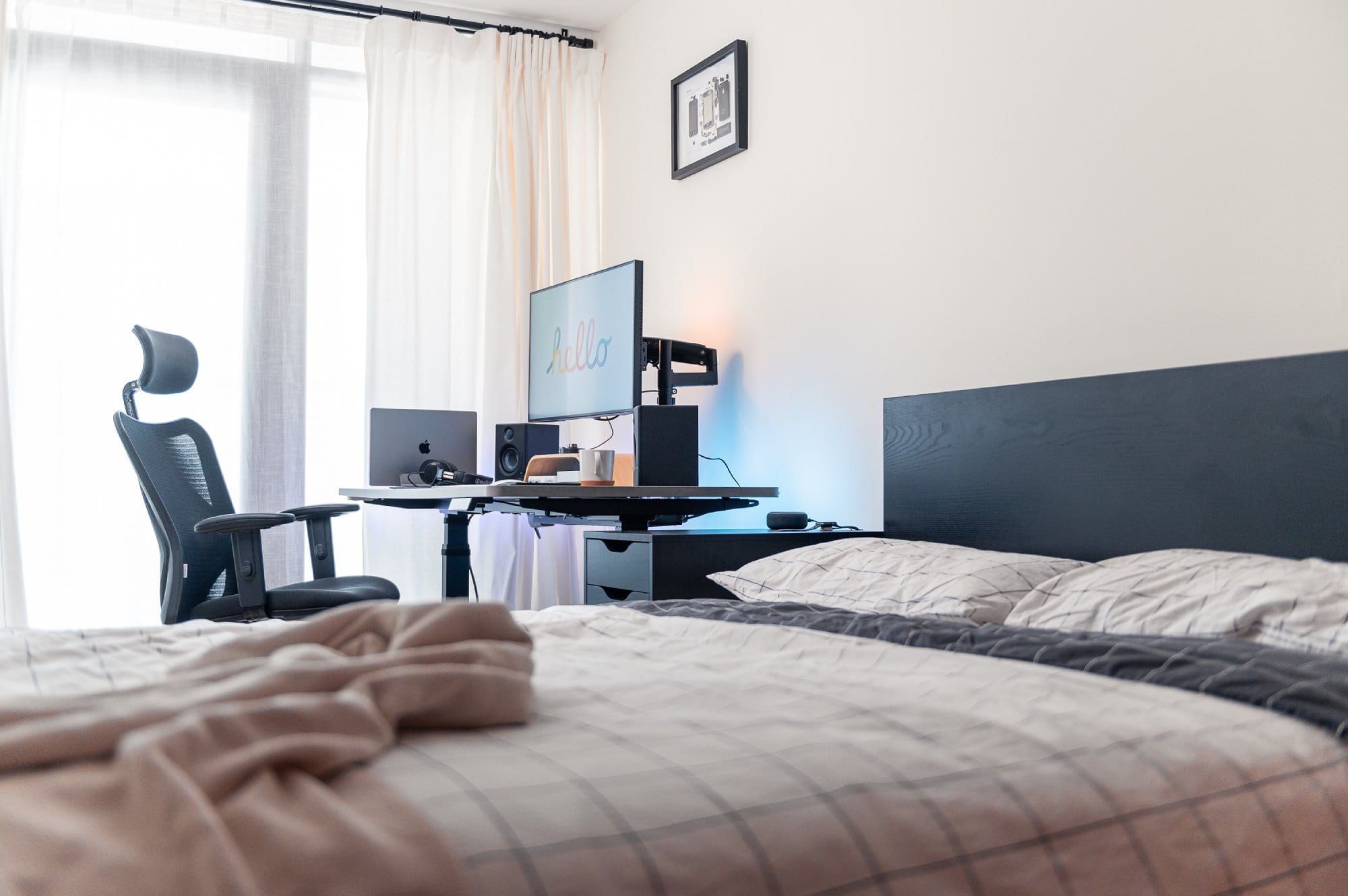 A bedroom with a bed in the foreground covered with a checkered duvet, looking towards a desk with an ergonomic chair and a monitor displaying “hello”, speakers, a laptop, and a framed picture on the wall