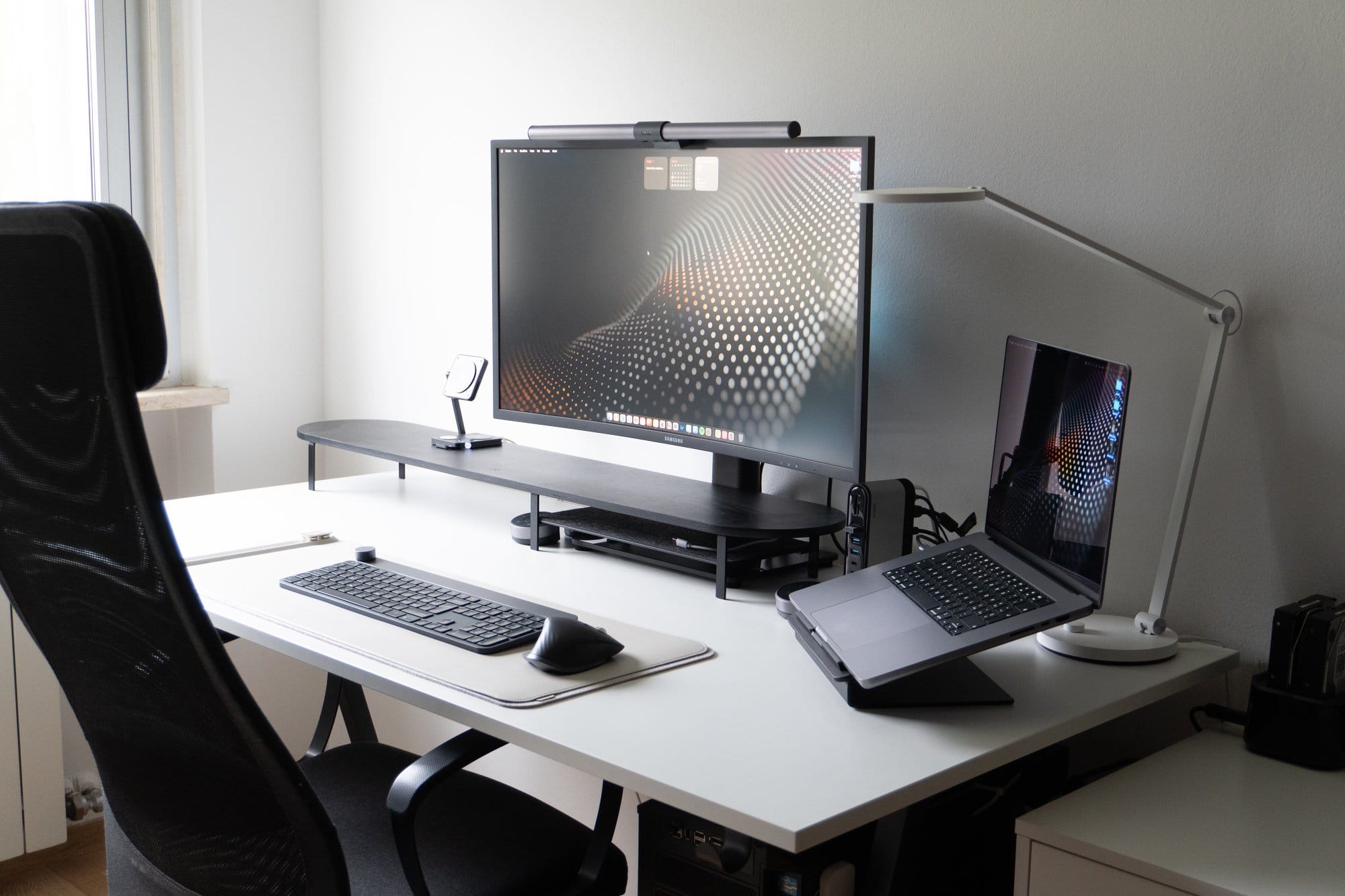A tidy workspace with a monitor, laptop on a stand, desk lamp, keyboard and mouse, set on a white desk beside an ergonomic chair