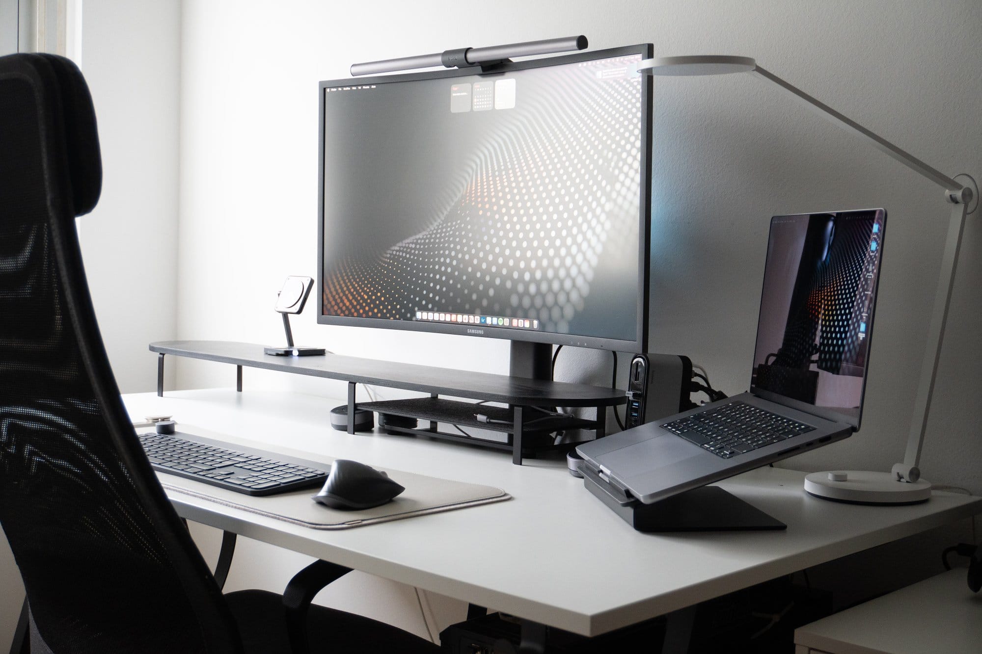A sleek home office setup with a monitor, a laptop on a riser, a keyboard, a mouse on a mat, and a desk lamp, arranged on a white desk with a black chair