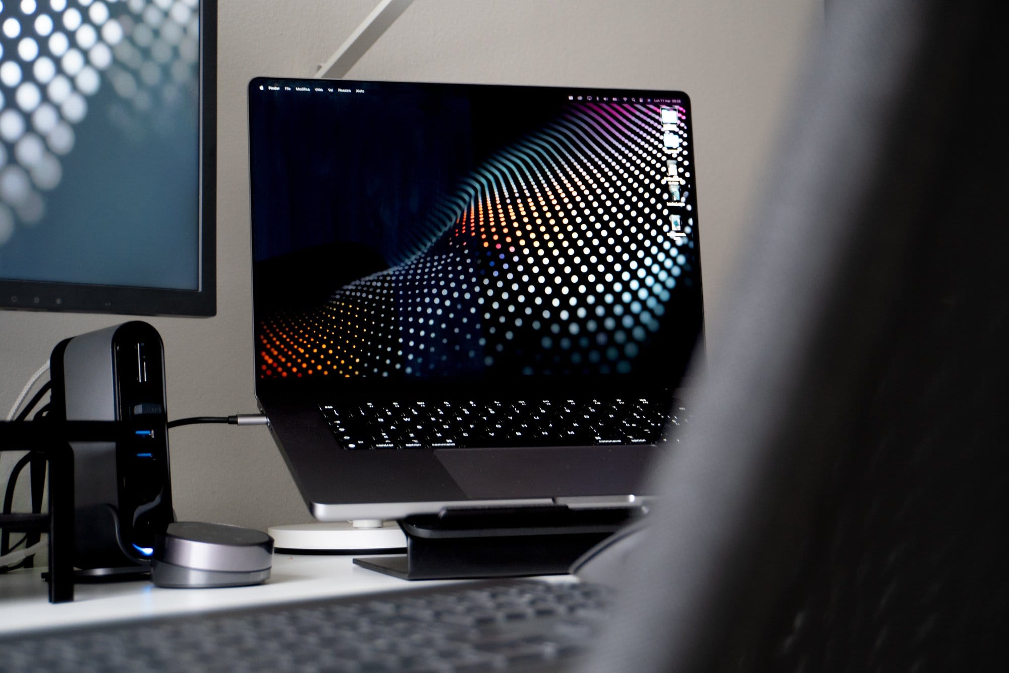 A close-up view of an Apple MacBook Pro on a stand displaying a colourful dot matrix design, with a blurred background featuring peripheral devices and part of a monitor