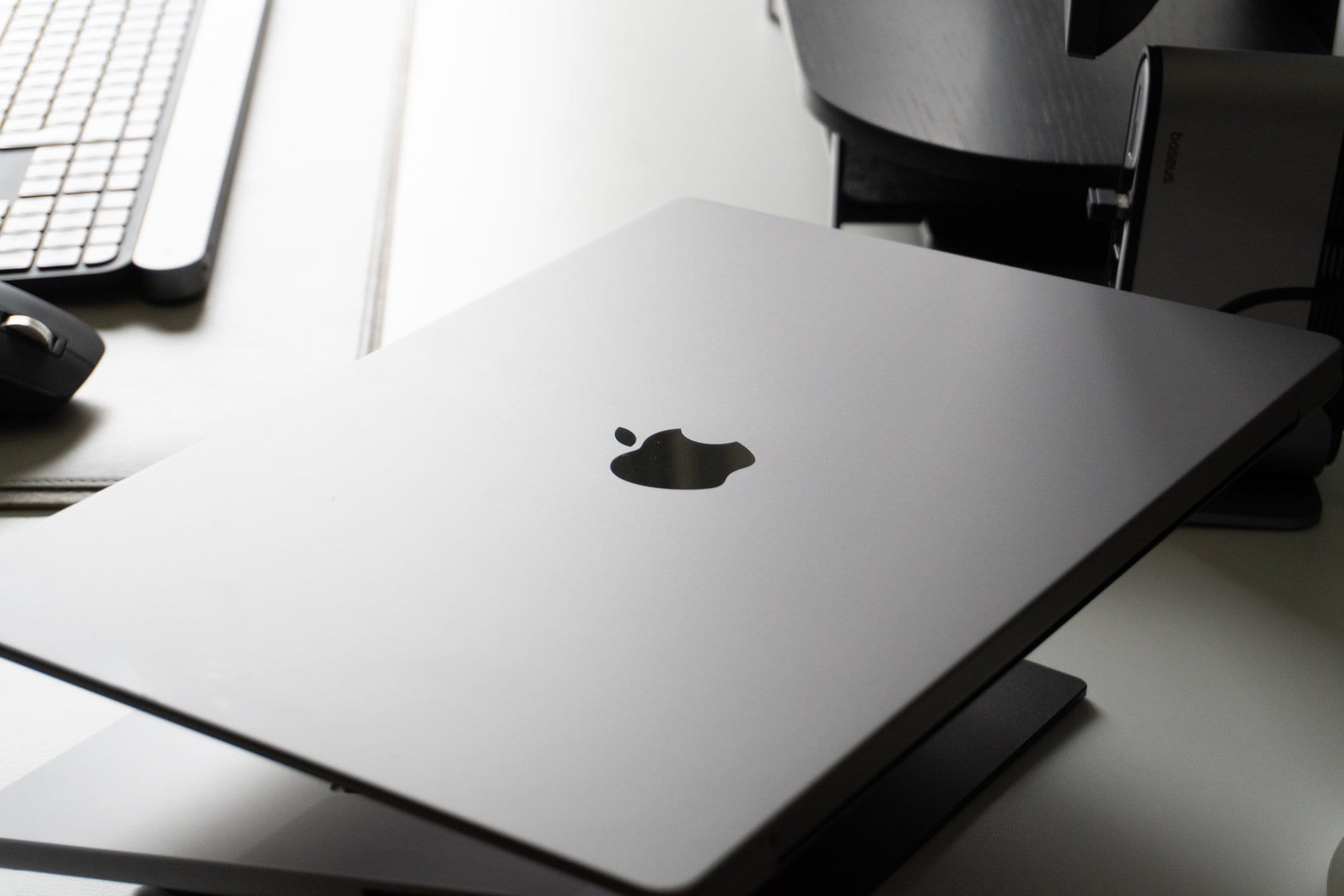 A closed Apple MacBook laptop on a stand, with a partial view of a keyboard and a mouse in the blurred background