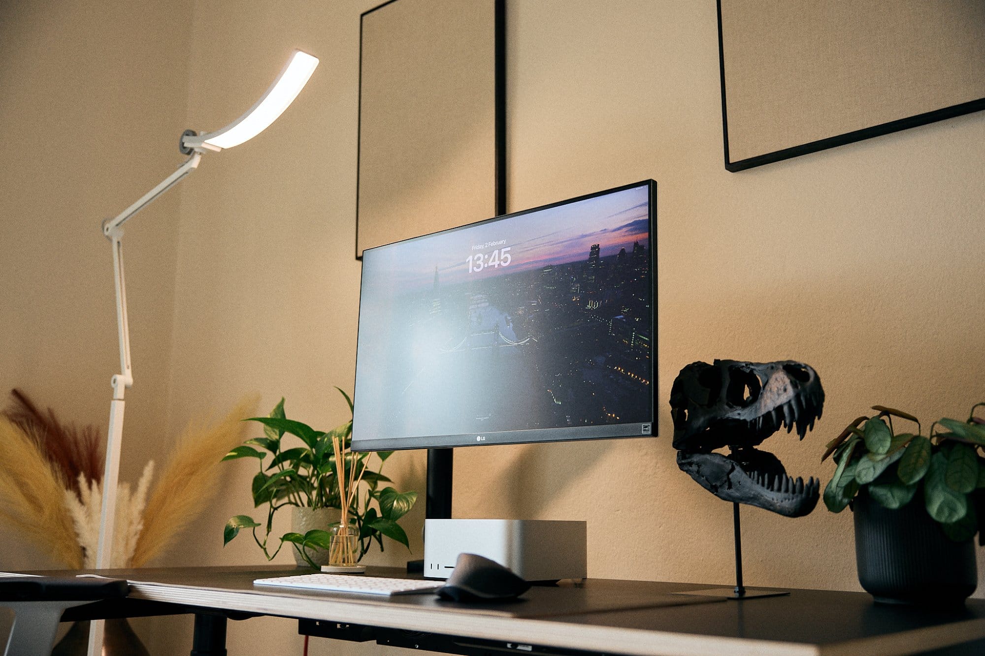 A modern workspace illuminated by a sleek desk lamp, featuring a monitor with a cityscape background, a dinosaur skull model, a green potted plant, and some dried pampas grass decoration