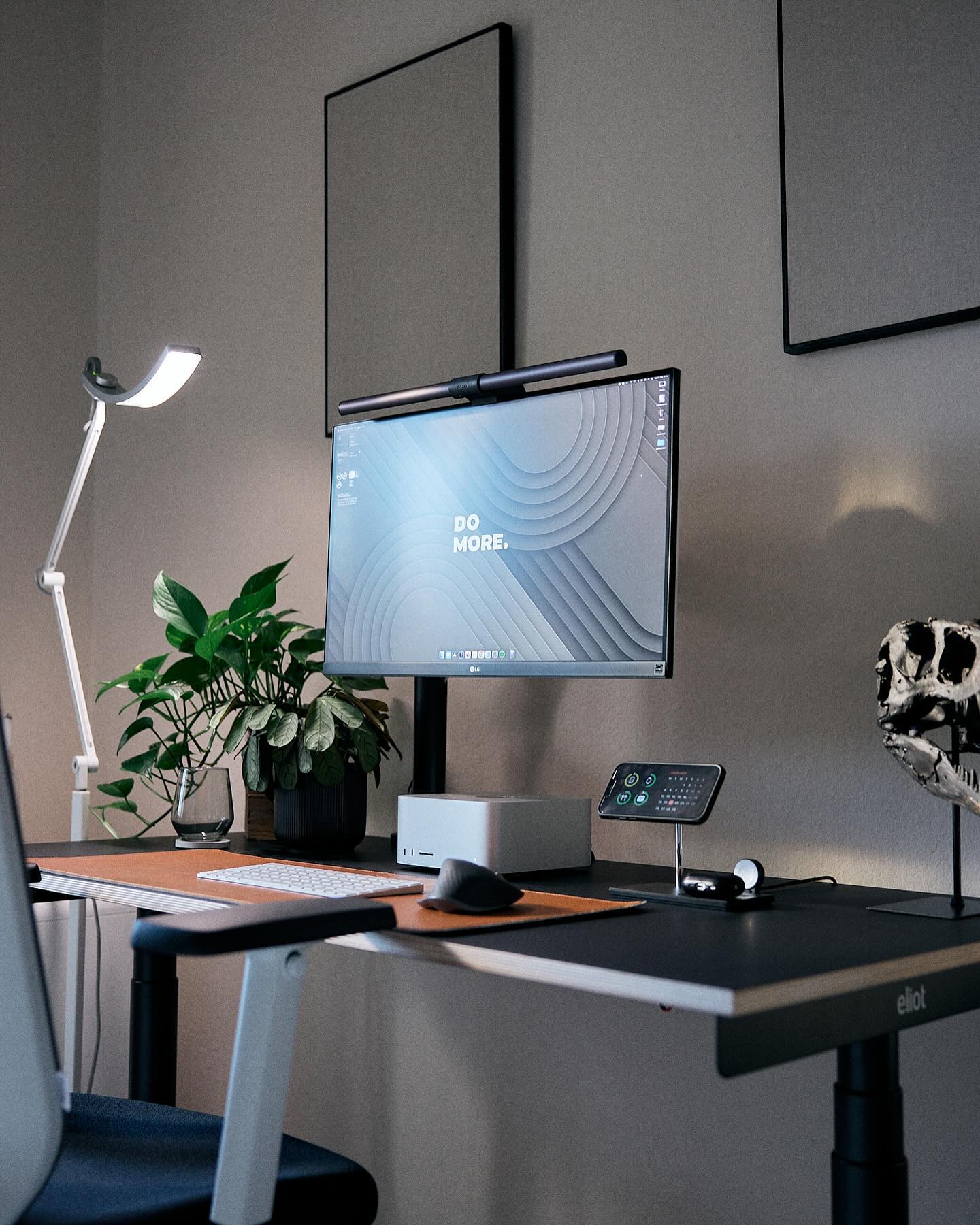 A well-organised work station with a height-adjustable desk, featuring a desktop computer with “DO MORE” on the screen, a soundbar, a phone on a stand, a Mac Studio, a decorative dinosaur skull, and green plants, all under a task lamp and against a wall with framed art