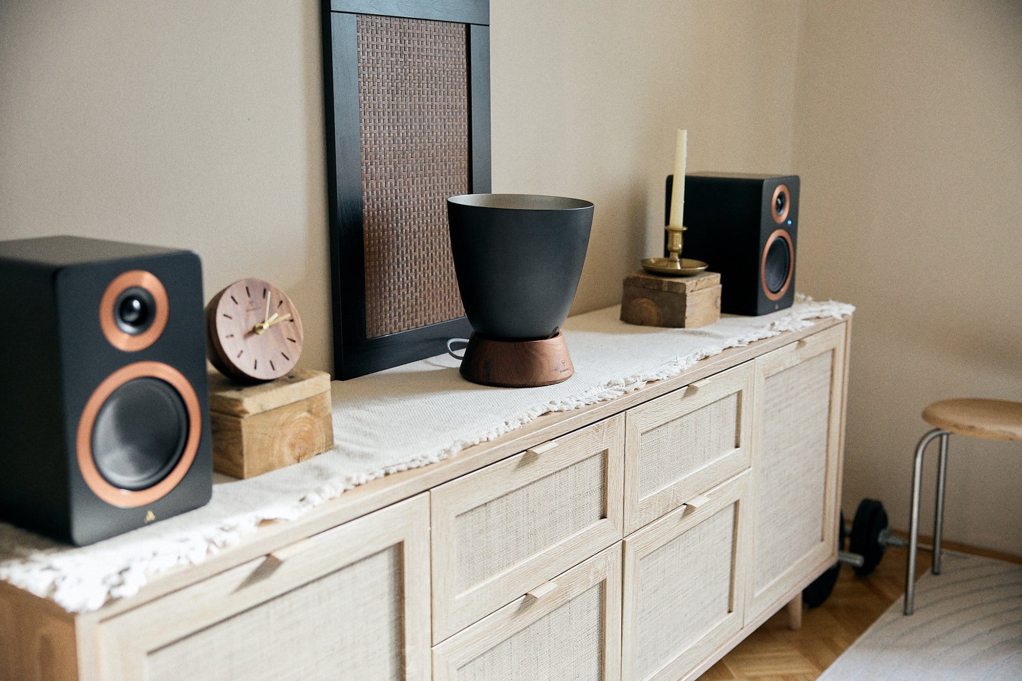 A stylishly arranged sideboard in a living room featuring bookshelf speakers, a modern vase on a wooden stand, a pink clock, a candle holder, and a woven runner