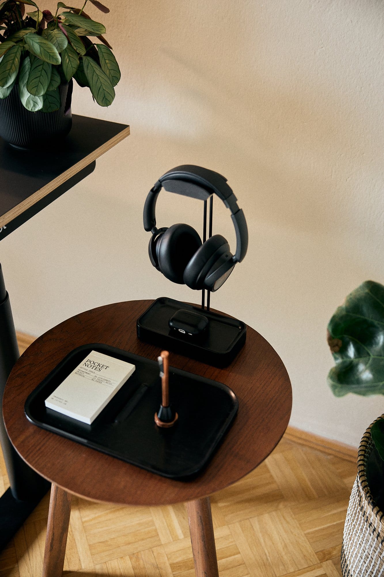  A contemporary side table holds a pair of headphones on a stand, a charging smartphone, and a pocket notebook beside a potted plant on a shelf above