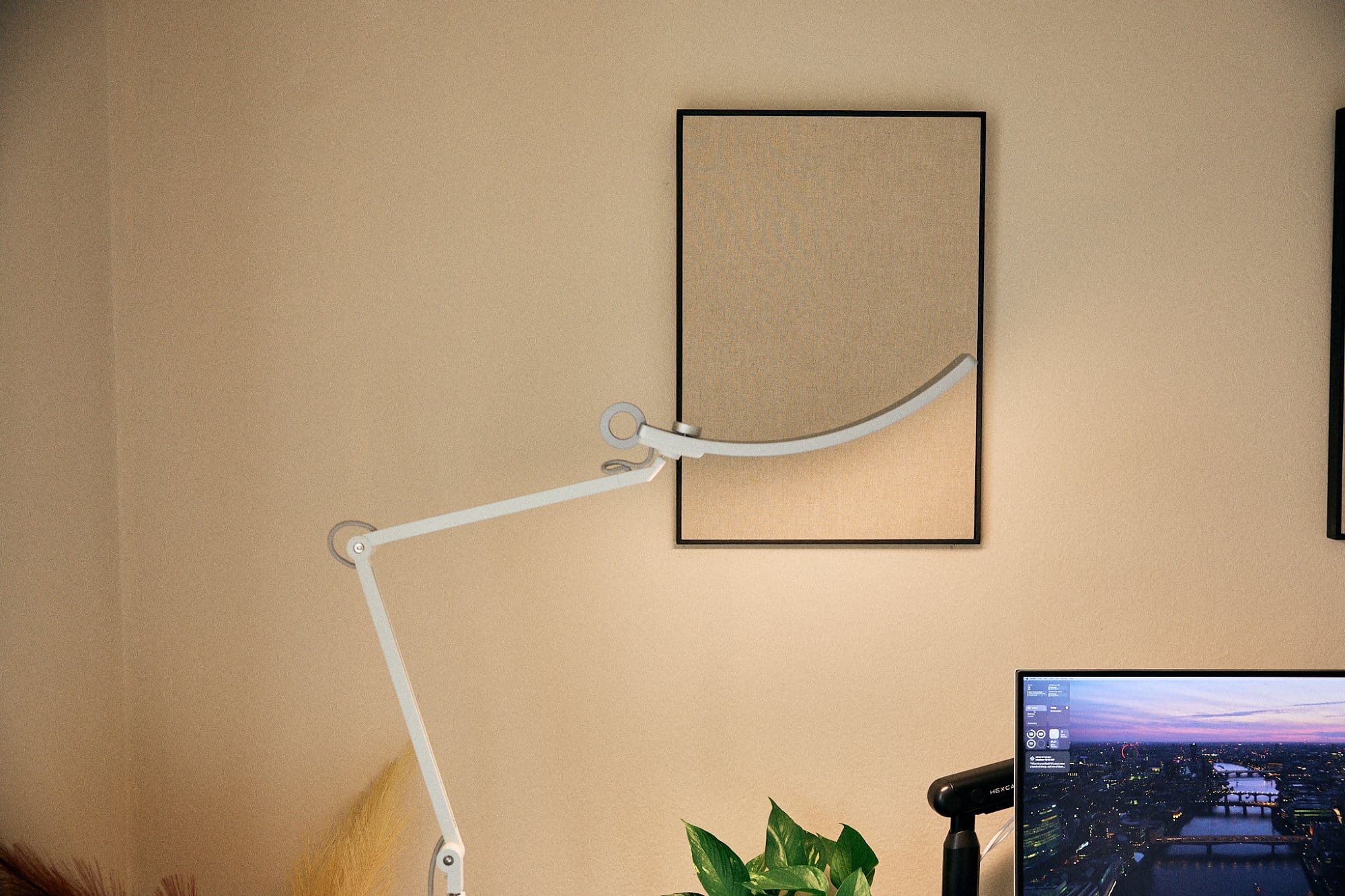 An adjustable white floor lamp is positioned next to a canvas wall art piece above a modern home office setup with a partial view of a monitor