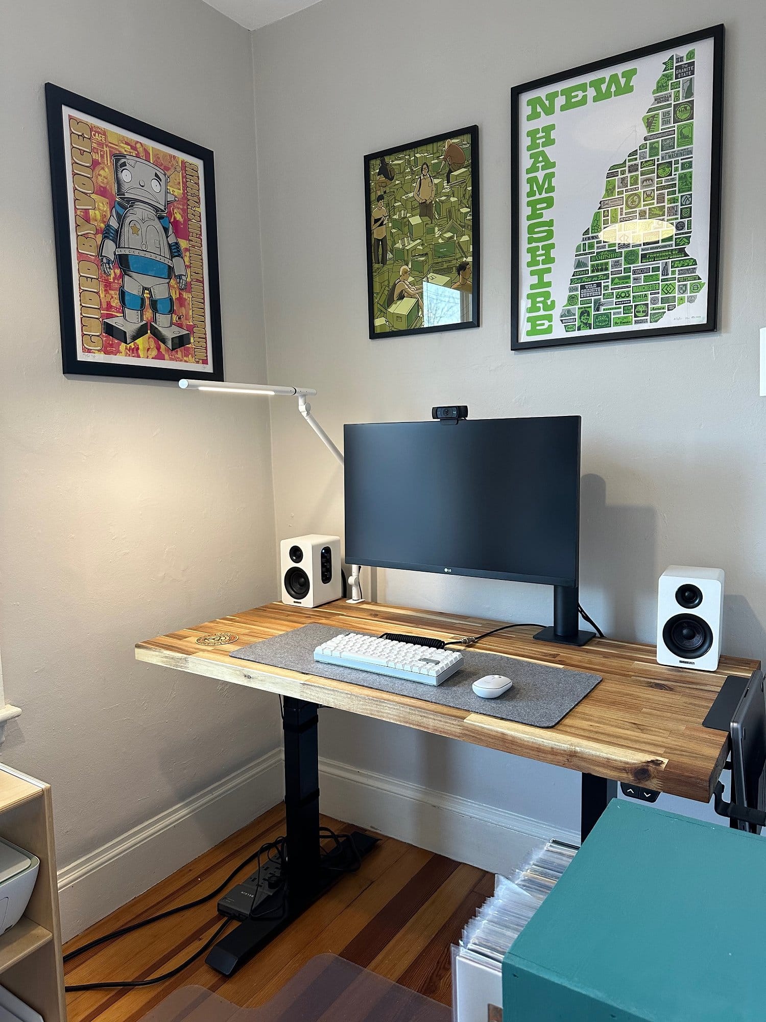 A home office corner with a height-adjustable desk holding a computer monitor and speakers, flanked by framed artwork on the walls