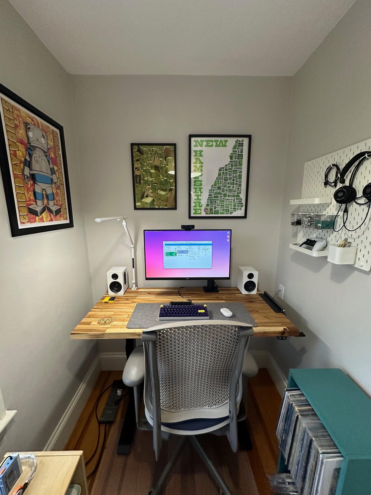 A small home office featuring a wooden desk with a computer, speakers, and various accessories, and decorated with framed artwork on the walls